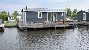 Ein Hausboot mit Deck, einer kleinen Veranda und einer anlegenden Mole, die sich in ruhiges Wasser erstreckt.