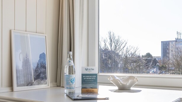 A hotel room with a large window, a framed picture, a bottle of water, a small vase, and a sign with text. - Calla - House Dünenblick - Calla (Sylt)