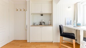 A compact kitchen with white cabinetry, a coffee maker, and a small countertop. There is a chair with a black cushion and a window with curtains. - Calla - House Dünenblick - Calla (Sylt)