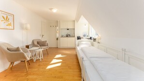 A modern, well-lit room with a sofa, a small table, a bed, and a kitchenette in the background. - Calla - House Dünenblick - Calla (Sylt)