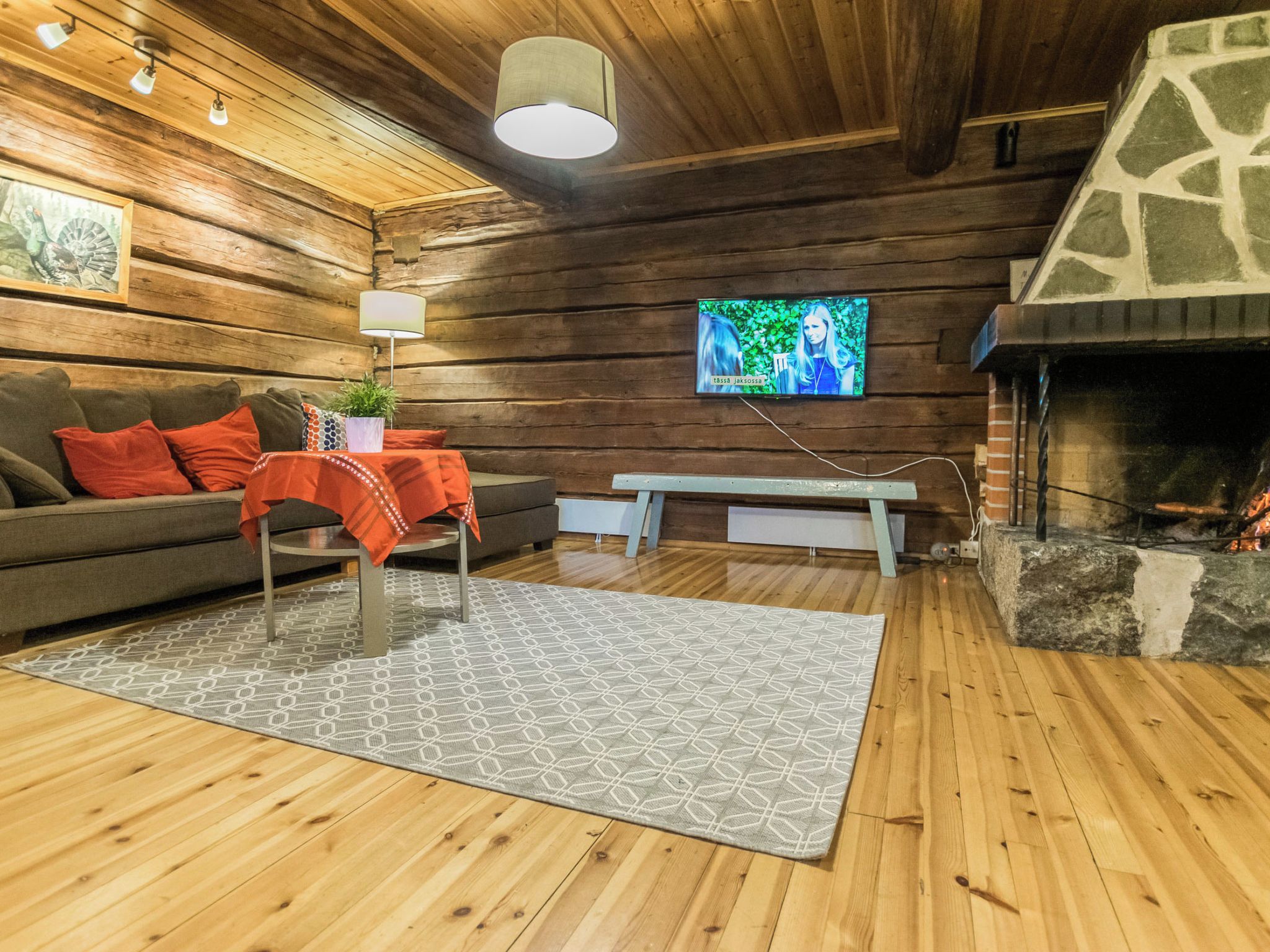 A cozy log cabin interior with a fireplace, a sofa with red cushions, a bench, and a television mounted on the wall.