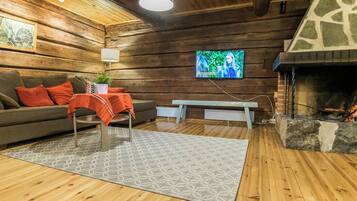 A cozy log cabin interior with a fireplace, a sofa with red cushions, a bench, and a television mounted on the wall.
