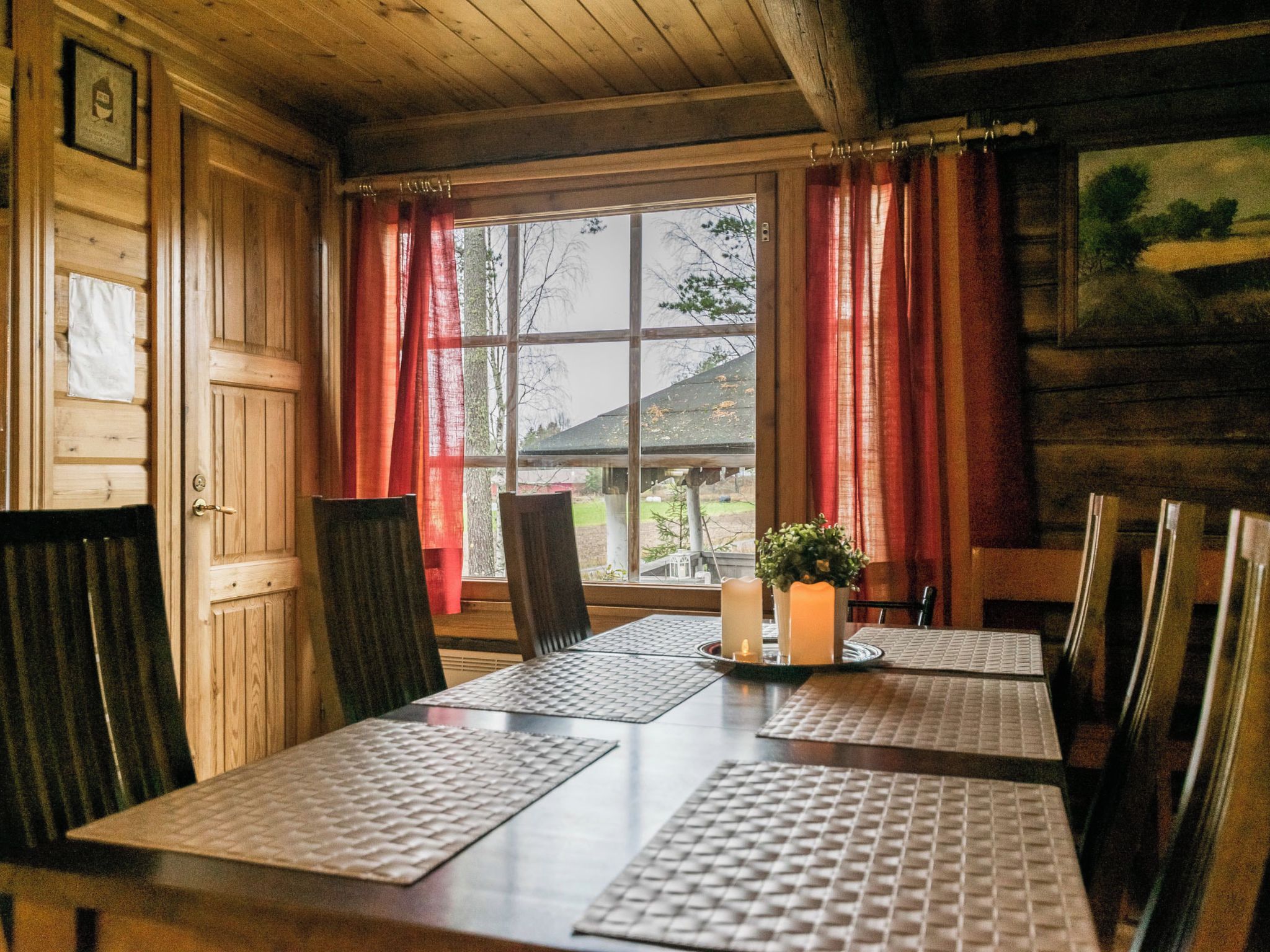 A wooden cabin interior with a dining table, chairs, a window with curtains, and a small plant on the table.
