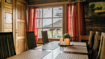 A wooden cabin interior with a dining table, chairs, a window with curtains, and a small plant on the table.