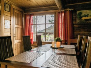 A wooden cabin interior with a dining table, chairs, a window with curtains, and a small plant on the table.