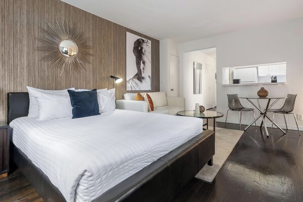 A modern hotel room with a large bed, a round mirror, a wall-mounted artwork, a small table with chairs, and a built-in TV.
