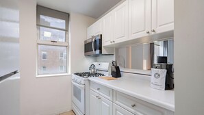Une cuisine compacte avec des meubles blancs, une cuisinière à gaz, un four à micro-ondes et une fenêtre donnant sur un immeuble en briques.