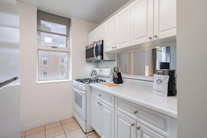 Una cocina compacta con muebles blancos, estufa a gas, microondas y ventana con vista a un edificio de ladrillo.