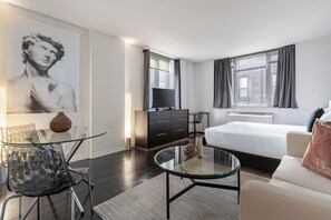 A modern hotel room with a glass coffee table, a sofa, a bed, a TV, and a large painting on the wall.
