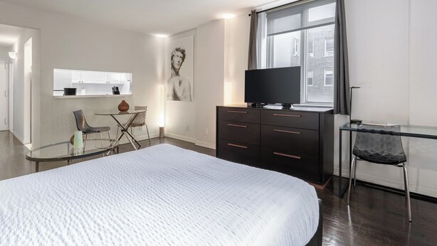 Une chambre moderne avec un grand lit, un téléviseur posé sur une commode, un bureau avec une chaise et une vue sur la ville par une grande fenêtre.