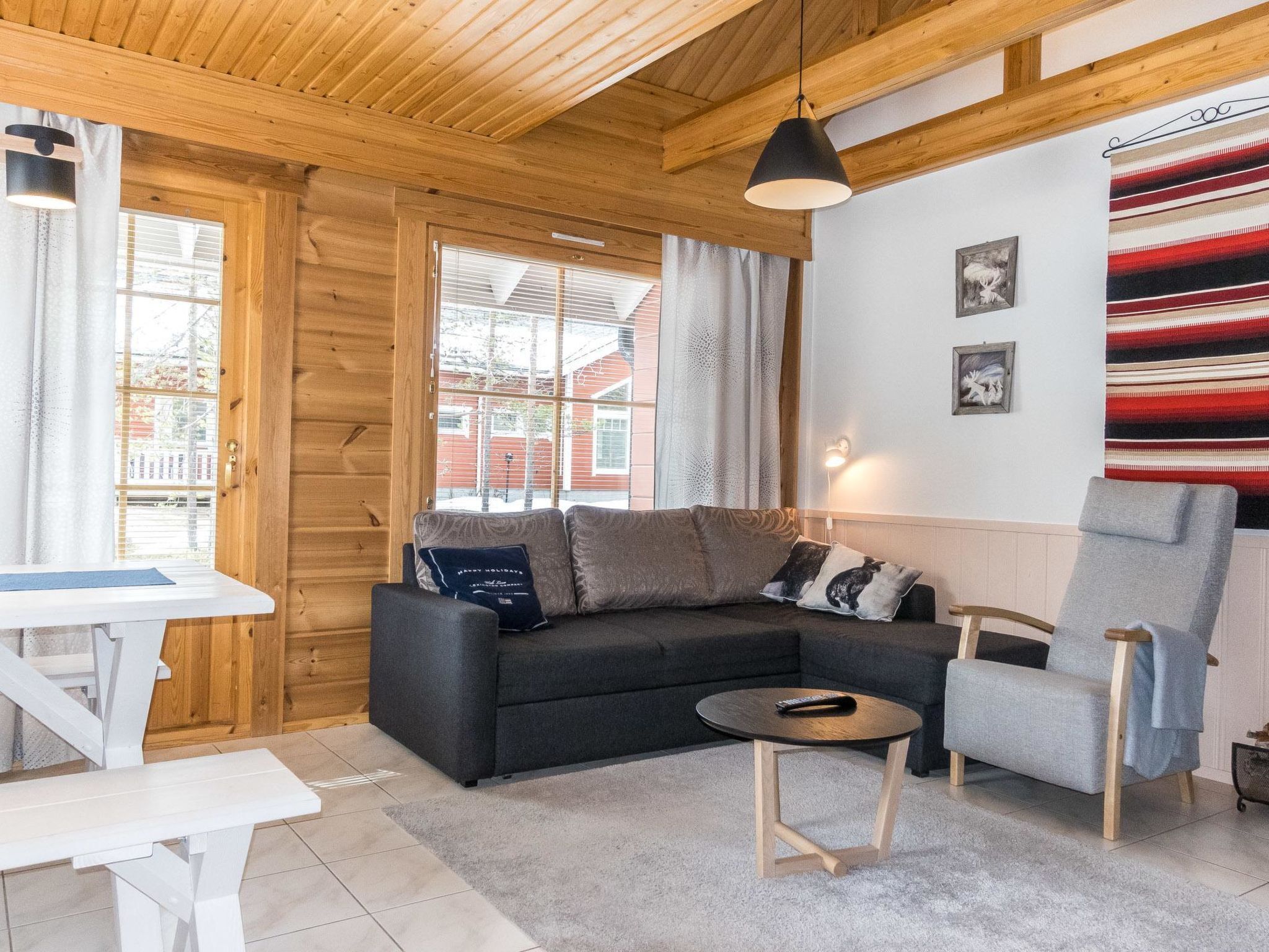 A cozy room with a wooden ceiling, a sofa, a chair, a small table, and a view of buildings outside.