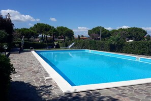 A rectangular swimming pool with clear blue water, surrounded by stone tiles and lush greenery.
