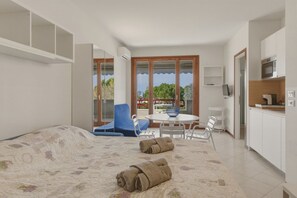A bedroom with a bed, a dining table with chairs, a kitchen area, and a view through a window.