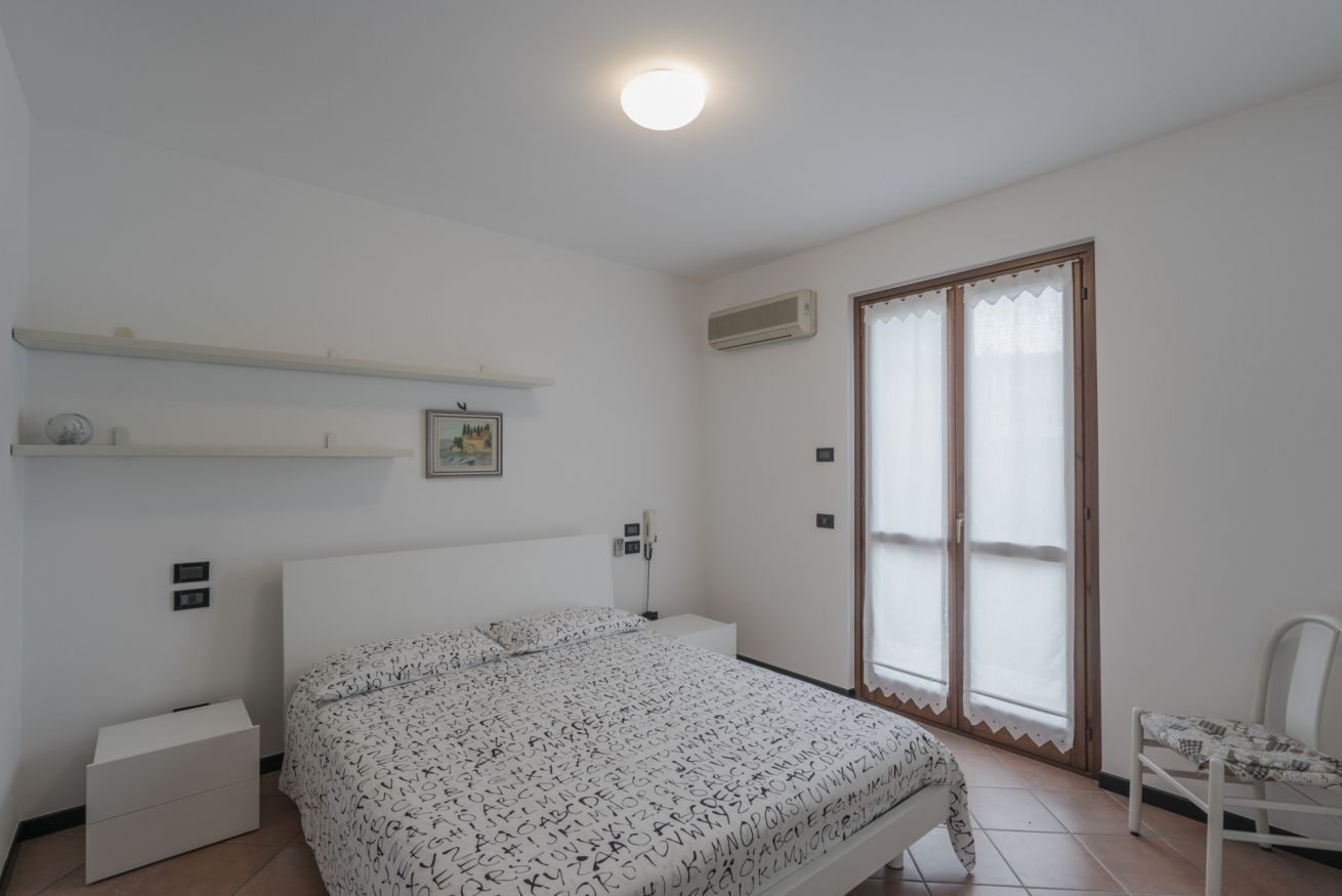 Une chambre à coucher comprenant un lit, une table de chevet, une chaise, une fenêtre avec des stores et un luminaire au plafond.