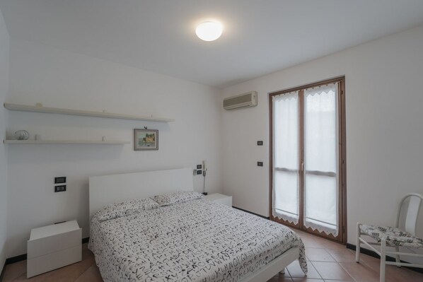 Un dormitorio con cama, mesita de noche, silla, ventana con persianas y luz de techo.