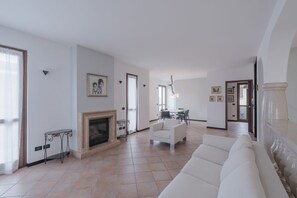 Een ruime woonkamer met een open haard, een witte bank en een eethoek met een tafel en stoelen.