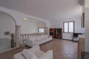 Amplia sala de estar con un sofá blanco, un sillón blanco más pequeño, una estantería de madera y un televisor.