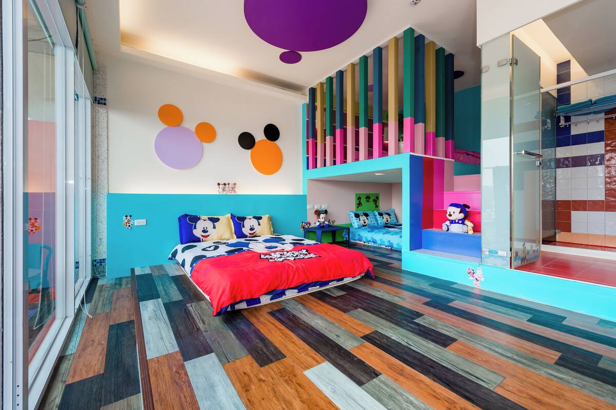 A modern bedroom with a colorful wooden floor, a bed with a red blanket, and a playful Mickey Mouse wall decoration.