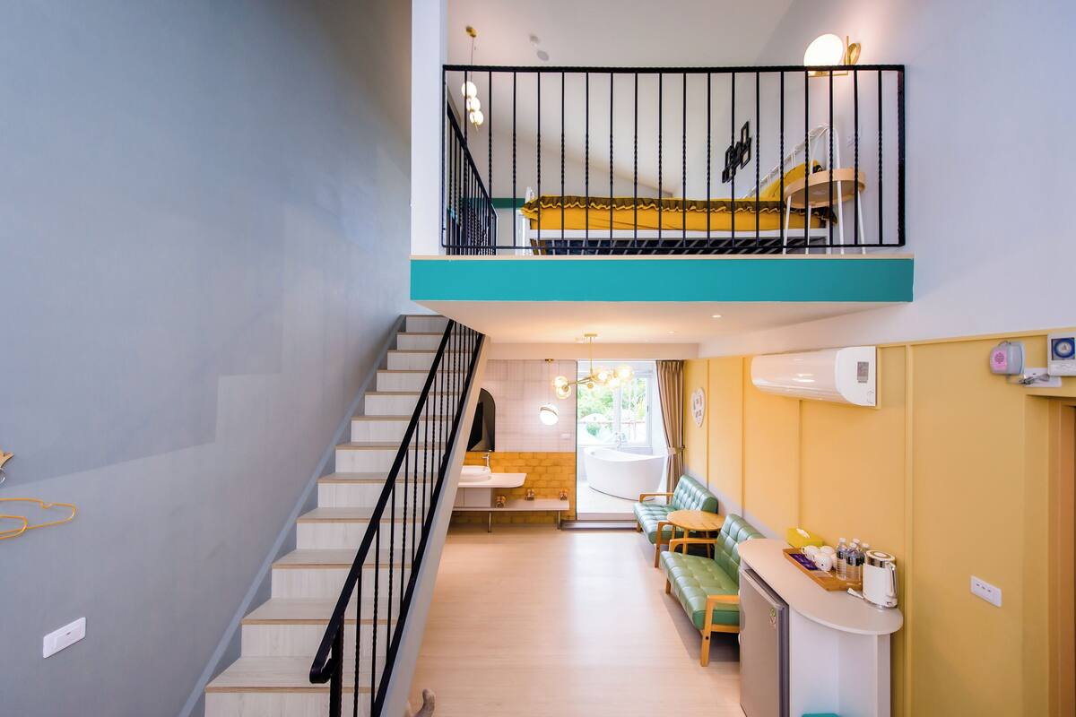 A modern hotel interior with a staircase leading to a balcony, a seating area with a small table, and a visible bathroom with a bathtub.
