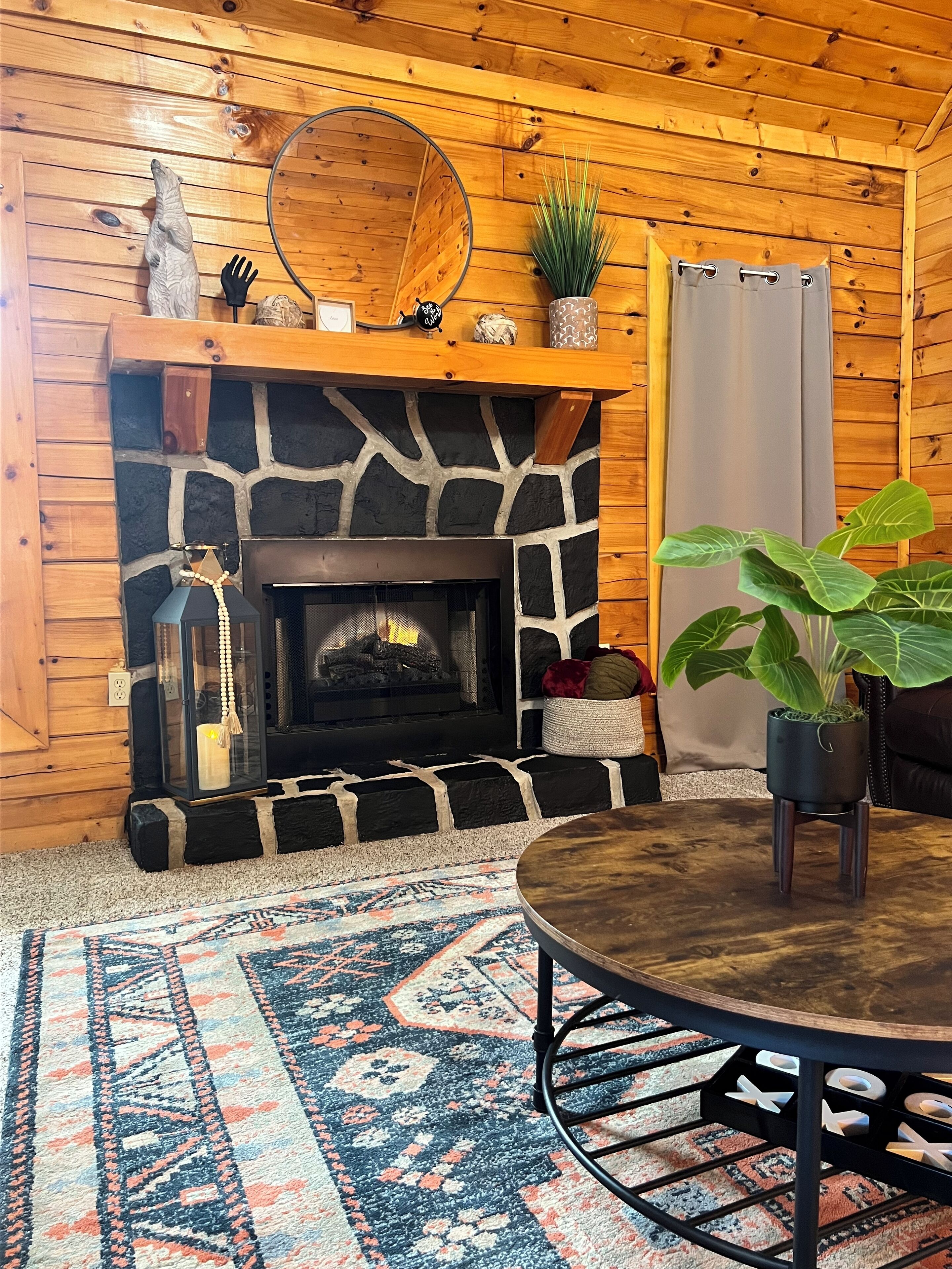 A cozy room with a stone fireplace, wooden walls, a round table, and a patterned rug.