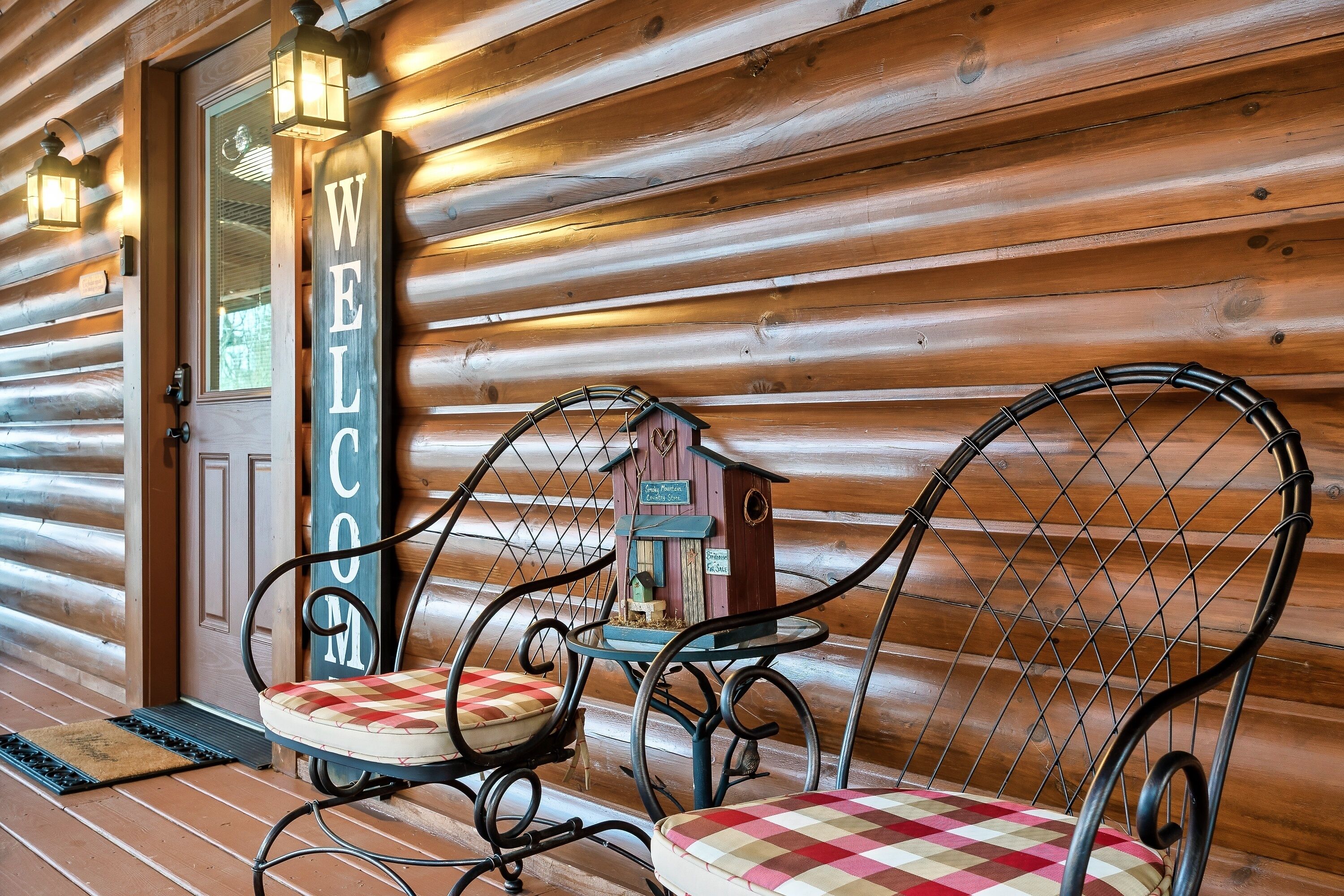 Eine Holzhütte mit einem 'WELCOME'-Schild, zwei Metallstühlen mit Karokissen und einem Vogelhäuschen auf einem Tisch.