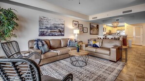 A modern living room with a sofa, coffee table, and chairs. There is a ceiling fan, a painting on the wall, and a kitchen area in the background.