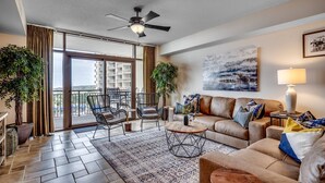 A spacious living room with a large sofa, two armchairs, a coffee table, and a ceiling fan. There are two potted plants, a large window with a view of buildings, and a painting on the wall.