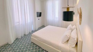Ein Hotelzimmer mit einem großen Bett, weißen Vorhängen, einem an der Wand befestigten Flachbildfernseher und einem gemusterten Boden.