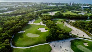 Luftaufnahme eines Golfplatzes mit Sandbunkern, Wasserhindernissen und einer Küstenlandschaft im Hintergrund.