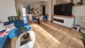 Ett modernt vardagsrum med en blå soffa, en matplats, en tv och ett kök i bakgrunden.