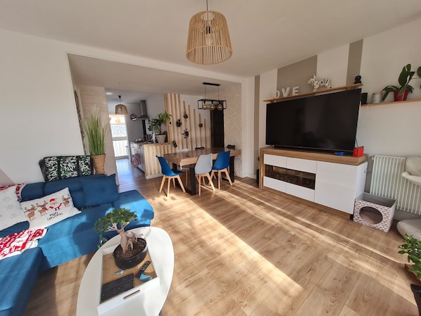 A modern living room with a blue sofa, a dining area, a TV, and a kitchen in the background.