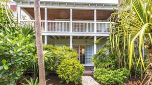 A two-story house with a covered porch, surrounded by a variety of tropical plants and a brick pathway leading to the entrance. - Luxury Sunset Key Cottage | Beach, Pool & Latitudes | 30+ Night Rental (Key West)