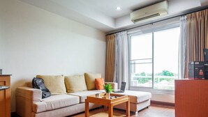 A modern living room with a sofa, a coffee table, and a laptop. There is a large window with curtains, an air conditioning unit, and a small desk with a computer. - Lake Side Condominium (Khon Kaen)
