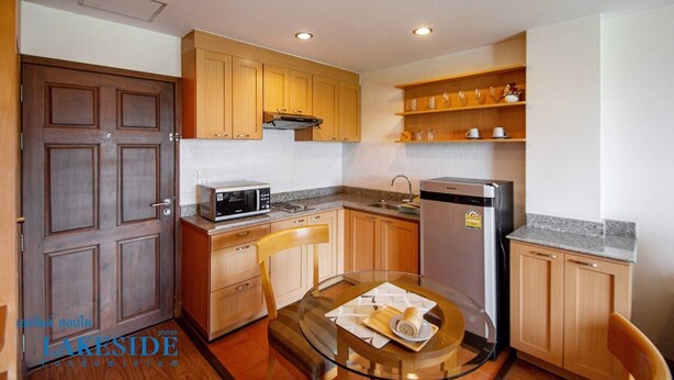 A compact kitchen with wooden cabinets, a microwave, a refrigerator, and a glass table with a napkin and a plate. - Lake Side Condominium (Khon Kaen)