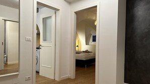 Un pasillo con lavadora, cama con almohadas y ventana con lámpara.