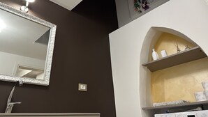 Un baño con un espejo grande, ducha, repisa con artículos de aseo y luz en el techo.