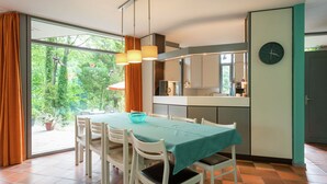 Un comedor con una mesa preparada para cuatro personas, una cocina visible a través de una puerta abierta y un reloj en la pared.
