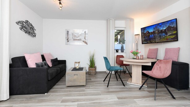 A modern living room with a black sofa, a round dining table, and a TV mounted on the wall. - Vacation apartment Seahorse (Esens)