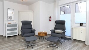 A modern living room with two grey armchairs, a small wooden side table, a white cabinet, and a large window with blinds. - Vacation apartment Seahorse (Esens)