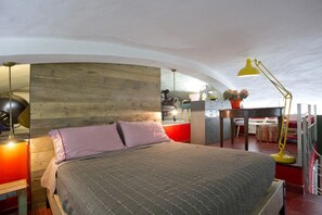 A bedroom with a bed, a wooden headboard, a desk with a chair, a yellow desk lamp, and a red wall. - LOFT n° 99 - Pavillion Italy (Siena)