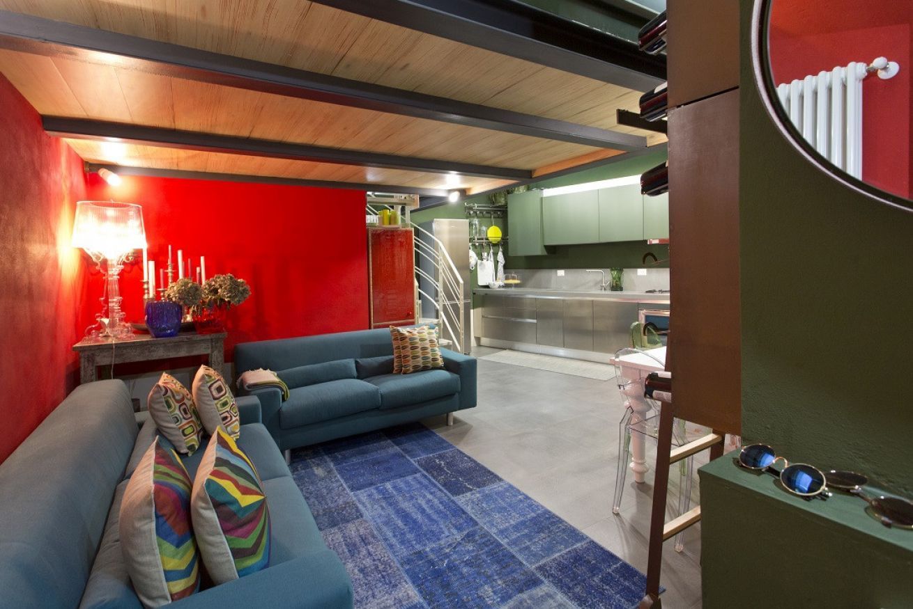 A modern living room with a blue sofa, a red accent wall, and a kitchen area in the background.