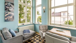 Une pièce avec un sol à carreaux, un canapé, une chaise, une table et une fenêtre donnant sur des immeubles et des arbres.