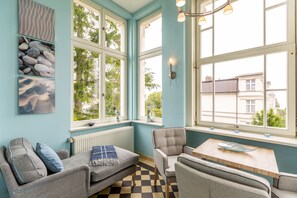 Une pièce avec un sol à carreaux, un canapé, une chaise, une table et une fenêtre donnant sur des immeubles et des arbres.