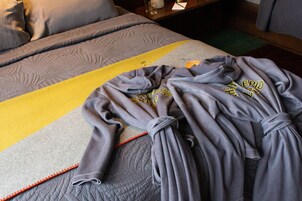 Una cama bien hecha con una colcha gris acolchada y una franja amarilla, una bata gris con texto en amarillo y una almohada gris.