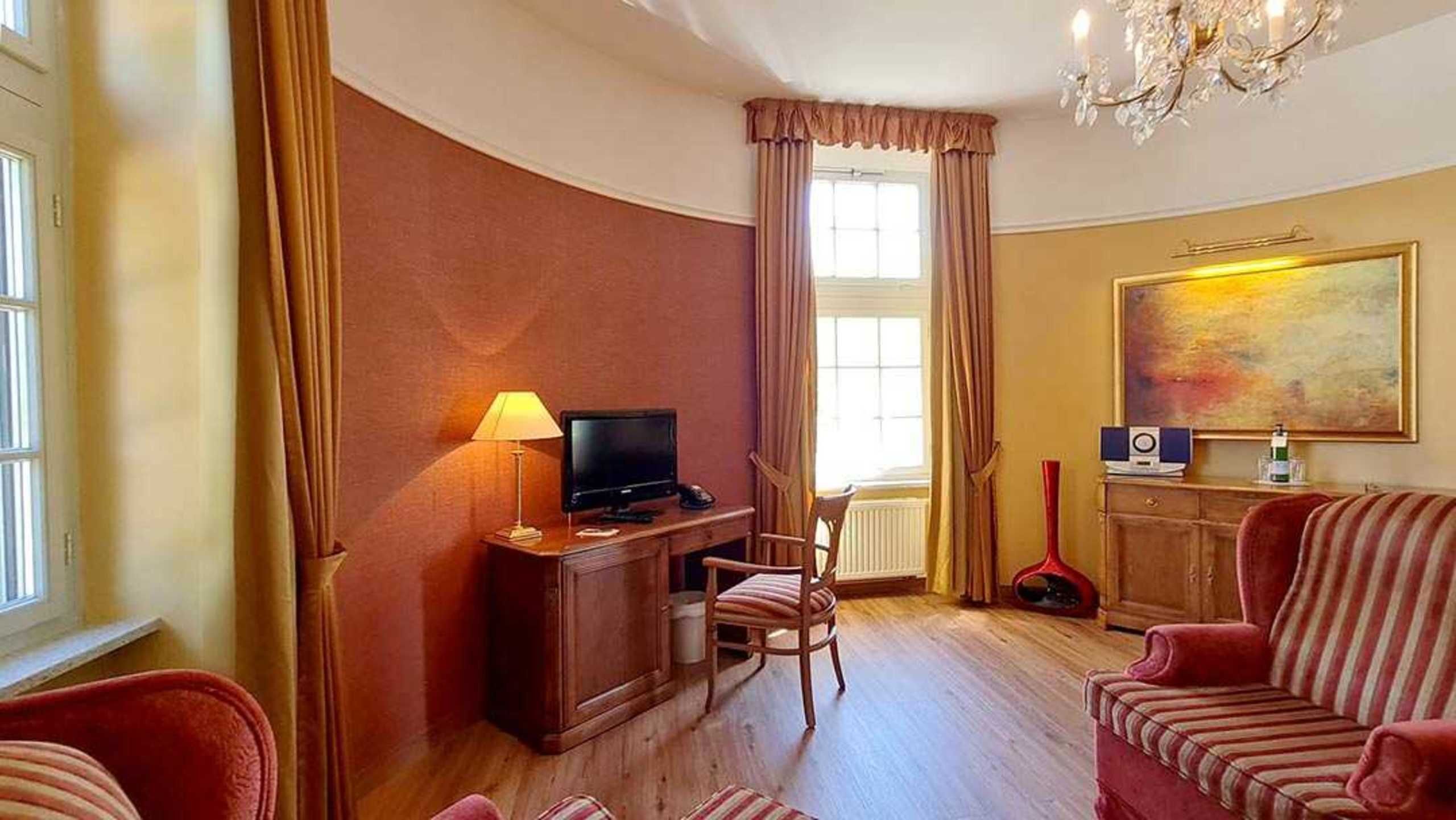 A hotel room with a wooden desk, a television, a red armchair, a striped sofa, and a painting on the wall.