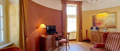A hotel room with a wooden desk, a television, a red armchair, a striped sofa, and a painting on the wall.