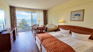 Ein Hotelzimmer mit Bett, Schreibtisch, Stuhl, Sofa und einem Balkon mit Meerblick.