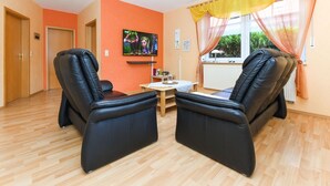 Una habitación con dos sillones de masaje negros, una mesa de madera, un televisor en la pared y una ventana con cortinas amarillas.
