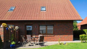 Une maison à deux étages, construite en briques, avec un toit recouvert de tuiles rouges, un porche à l’avant avec une table et des chaises, et une petite fenêtre au deuxième étage.