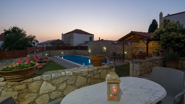 A poolside area with stone walls, a pool, lounge chairs, and a table with a lantern. - Skandalis Apartments with sea & pool view (Klio) (CHORAFAKIA CHANIA)